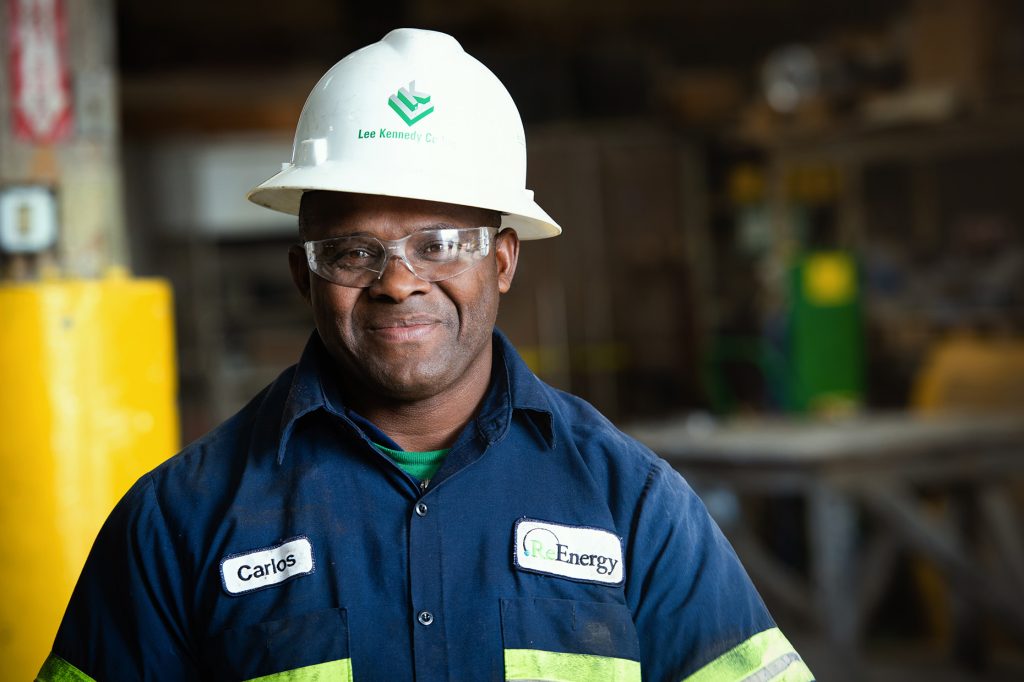 A man named Carlos stands confidently in a ReEnergy uniform and hard hat inside a recycling facility.